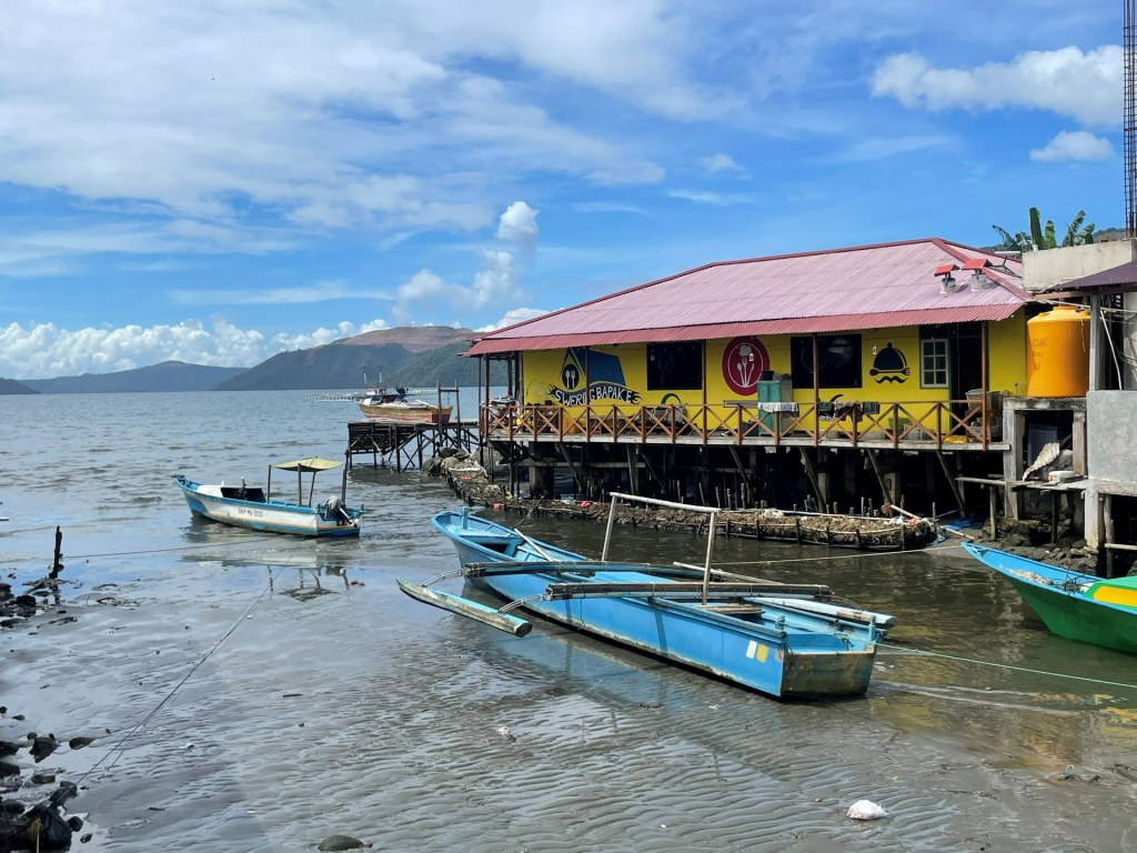 Perahu-perahu nelayan di pesisir Teluk Buli saat air surut, dengan rumah panggung dan bukit di kejauhan